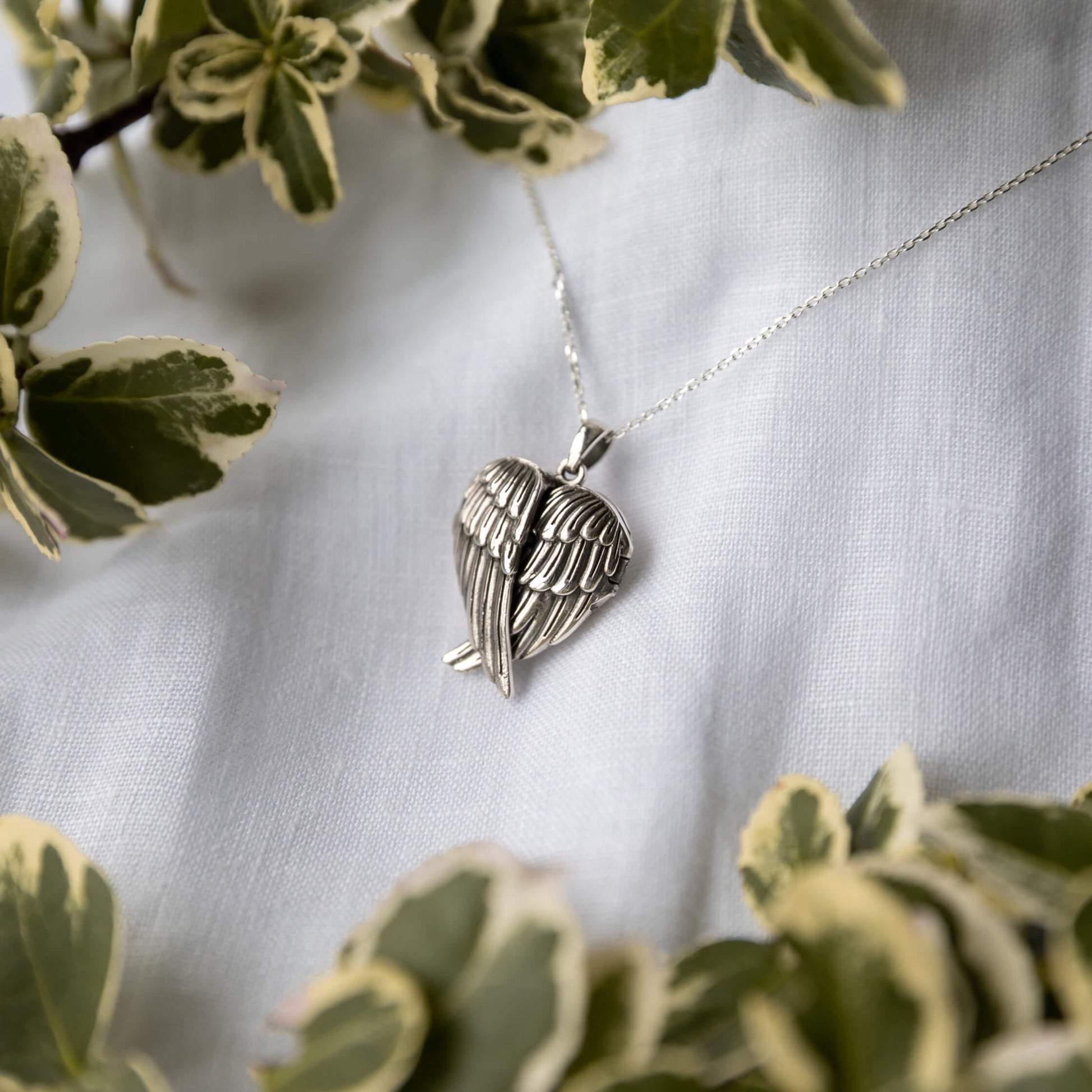 Silver wing-shaped pendant necklace on a white fabric background with green leaves.