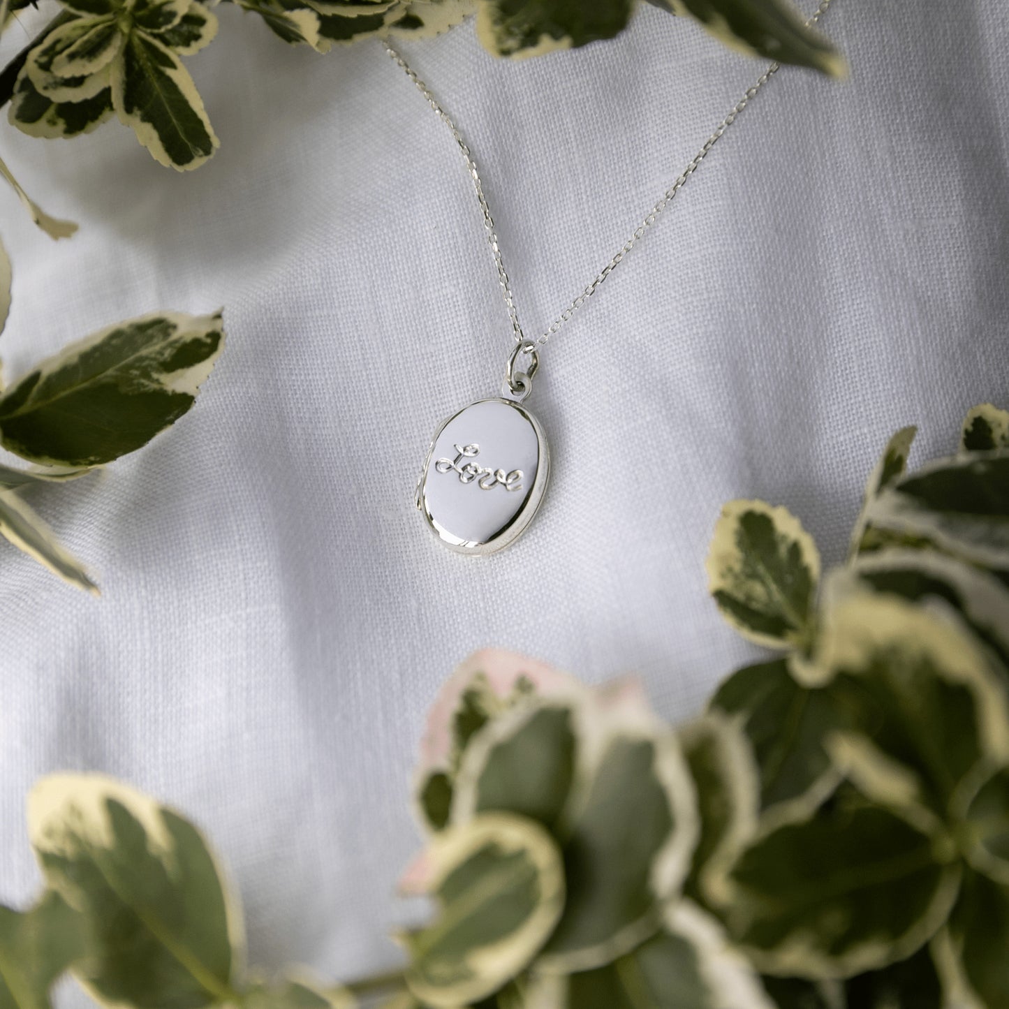 Silver necklace with a locket on a white fabric background surrounded by green leaves.