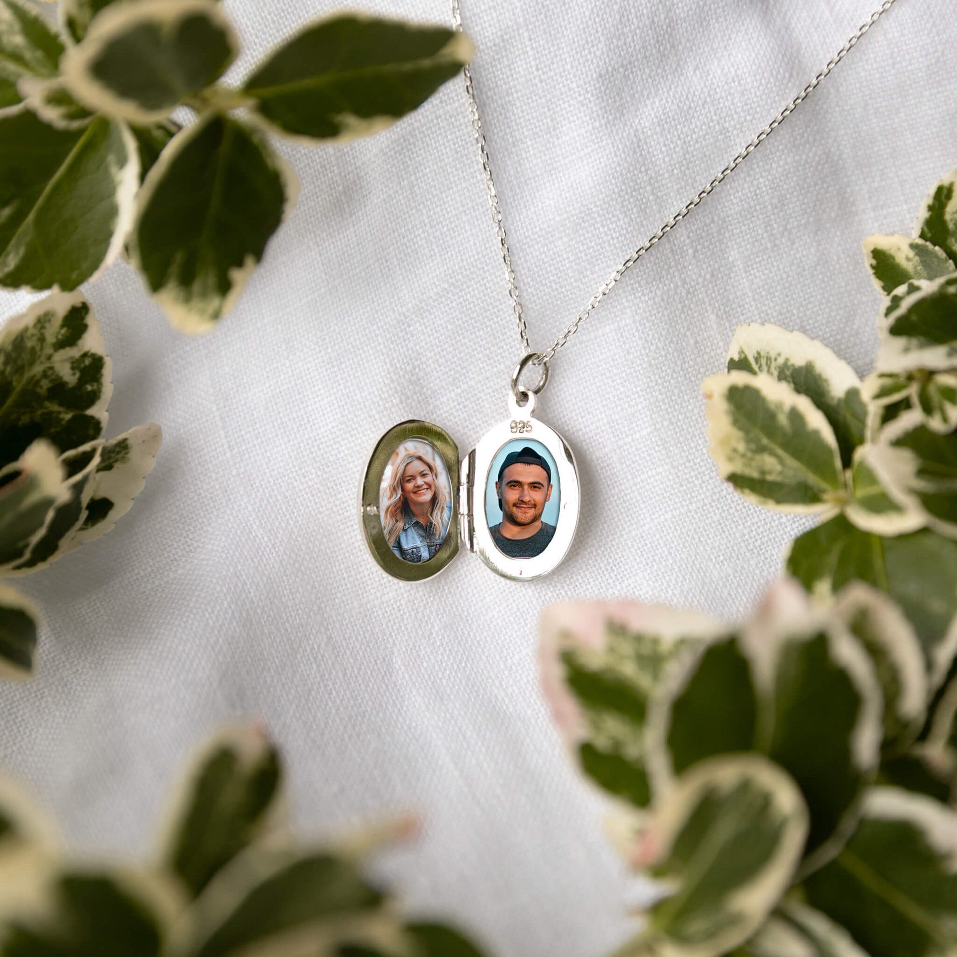 Silver locket with two photos on a white fabric background with green leaves.