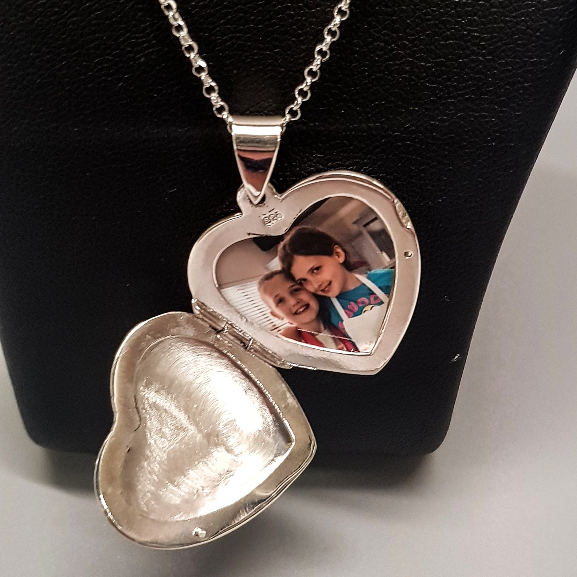Large silver heart shaped photo locket necklace on silver chain displayed on a black bust. Locket is open showing photo of two little girls inside.